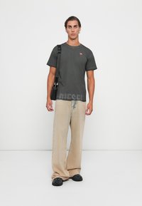 Graues kurzärmliges T-Shirt mit "DIESEL" in großen Buchstaben bedruckt, kombiniert mit beigen weiten Hosen. Das Model hält eine schwarze Crossbody-Tasche.