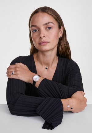 Maglietta nera a maniche lunghe con pieghe. Gli accessori includono orecchini circolari, un orologio d'argento, una collana e bracciali con accenti neri e argentati.