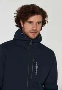 Hombre con cabello rizado que lleva una sudadera con cremallera de color azul marino y un cuello alto, con el logo de "Sail Racing" en el bolsillo del pecho.
