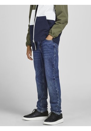 Person wearing blue denim jeans, black sneakers with white soles, and a green, white, and navy zip-up jacket with one hand in pocket.