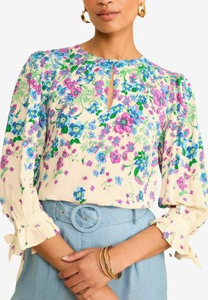 Femme portant un chemisier crème à fleurs roses, bleues et vertes, manches bouffantes avec nœuds à volants, rentré dans un pantalon ceinturé bleu clair.