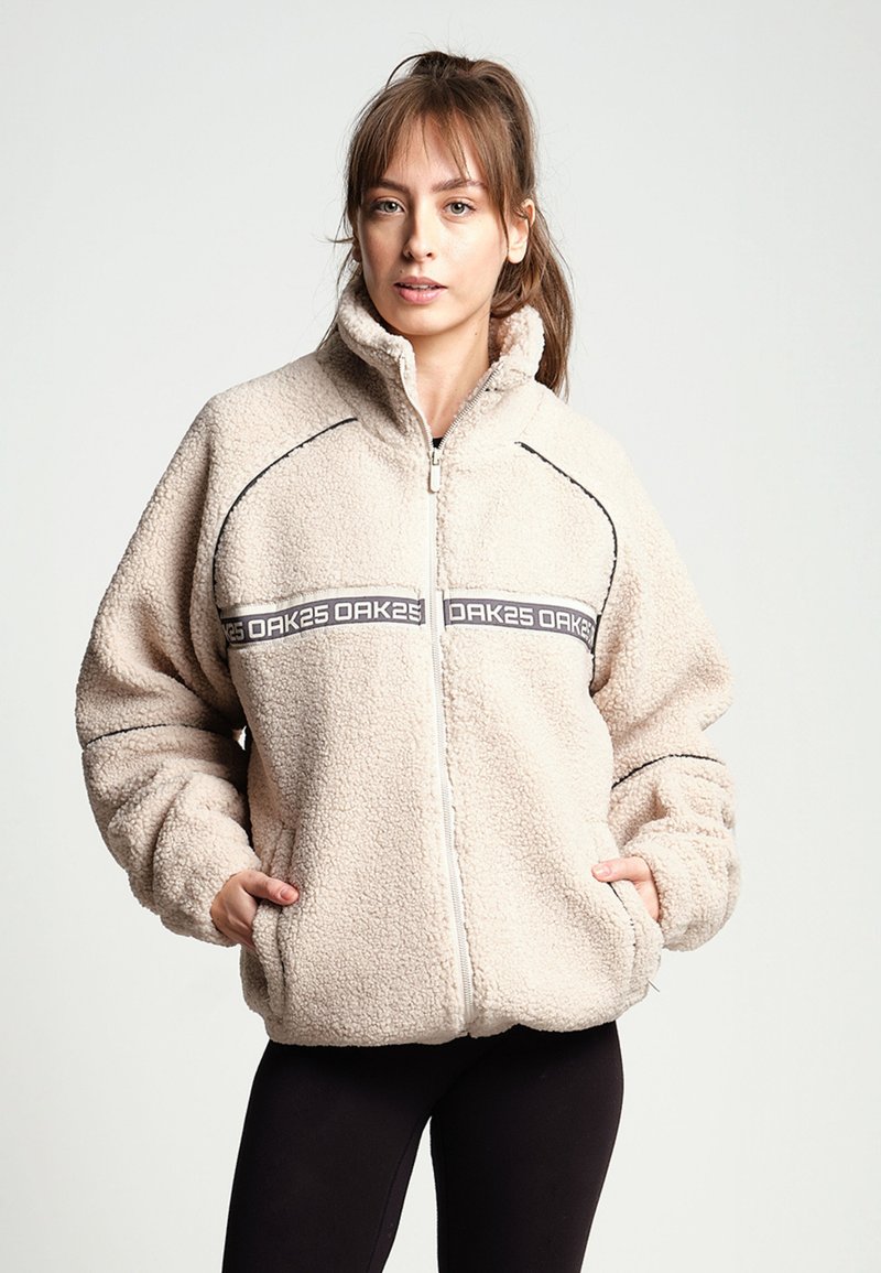 Giacca a zip in pile beige con colletto alto, decorata con accentuazioni nere e una fascia con logo sul petto. Presenta tasche laterali.