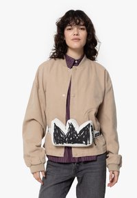 Beige corduroy jas met een drukknoopsluiting, gecombineerd met een gestreept shirt en donkere spijkerbroek. Heeft een witte heuptas met zwart bergontwerp.