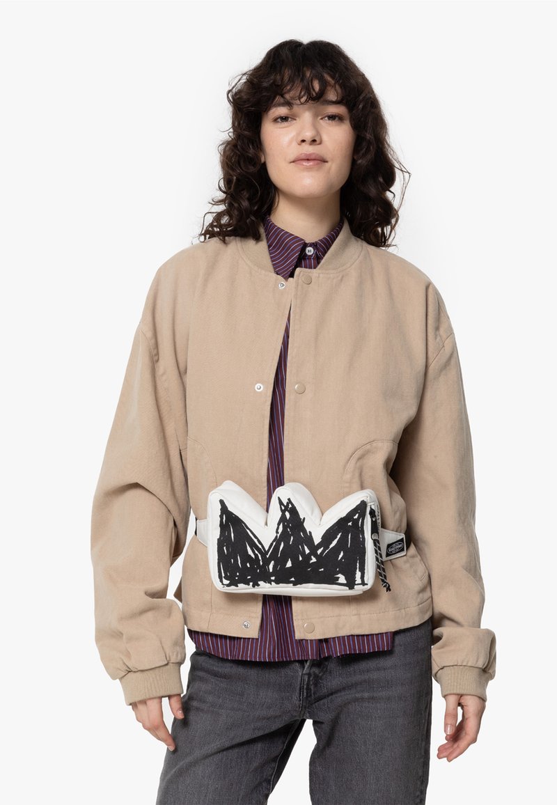 Beige corduroy jas met een drukknoopsluiting, gecombineerd met een gestreept shirt en donkere spijkerbroek. Heeft een witte heuptas met zwart bergontwerp.