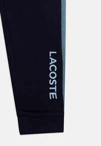 Tamno plave trenirke s svijetloplavim bočnim prugama i bijelim natpisom "LACOSTE" na vanjskom dijelu nogavice. Izrađene su od mekanog, rastezljivog materijala.