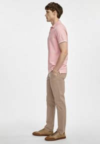 Jasnoróżowa koszula polo z białymi dodatkami, w zestawieniu z beżowymi spodniami i brązowymi trampkami. Model stoi bokiem, z rękami w kieszeniach, na neutralnym tle.