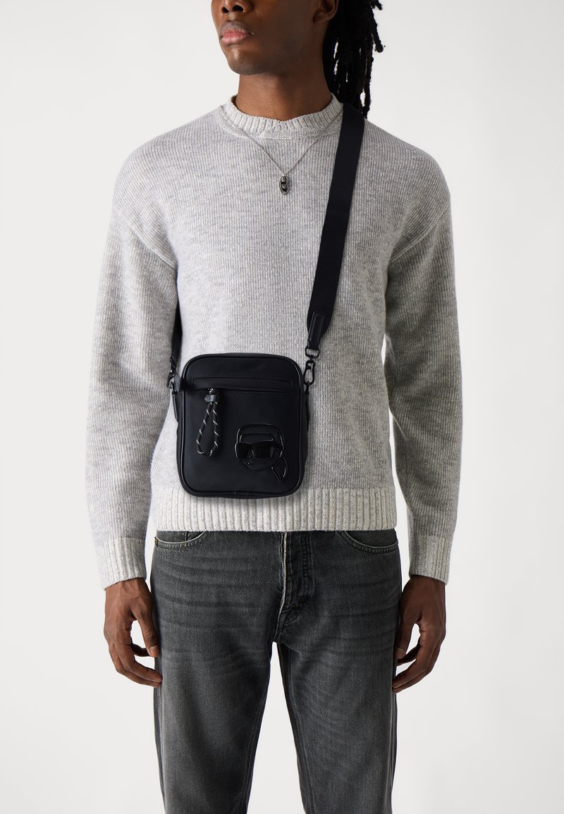Black crossbody bag made of textured material, featuring a zippered pocket and a braided accent. Adjustable strap, square shape.