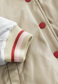 Beige jacka med en vit ärm, som har en ribbad mudd i beige, rött och rosa ränder. Röda tryckknappar framtill. Slät textur.