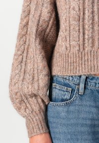 Beżowy sweter w warkoczowy splot z grubą teksturą, z bufkami na rękawach i prążkowanym dołem, w zestawie z jasnoniebieskimi dżinsem.