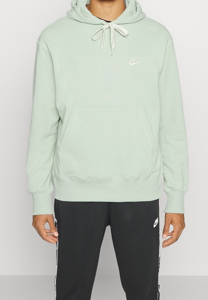 Světlězelená mikina Nike s kapsou na přední straně, kapucí na šňůrku a vyšitým logem. Detail žebrovaných manžet a lemu. V kombinaci s tmavými kalhotami.