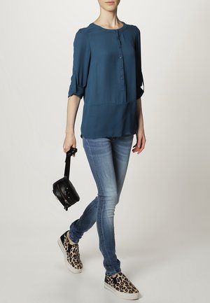 Blusa azul verdoso con botones, mangas remangadas, combinada con vaqueros pitillo azules. Se lleva con zapatos slip-on estampados de leopardo y un bolso pequeño negro.