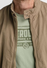 Khaki jacket with a zippered front, standing collar, and smooth texture, layered over a pale green T-shirt featuring a graphic print.