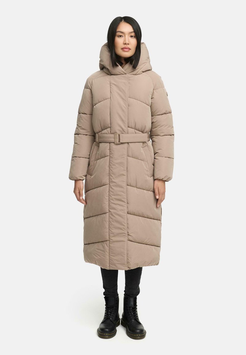 Lange beige pufferjas met een capuchon, een ceintuur in de taille en een diagonale gestikte design. Gekleed over zwarte jeans en zwarte veterschoenen.