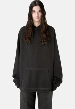 Donna con capelli lunghi e scuri che indossa una felpa oversize nera e pantaloni neri abbinati su uno sfondo chiaro e semplice.