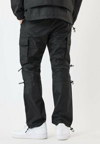 Zwarte cargo-broek met verstelbare boorden en zijzakken, gemaakt van een glad, lichtgewicht materiaal. Gepaard met witte sneakers.