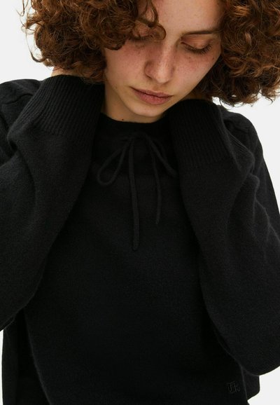 Mujer con cabello rizado que lleva un suéter negro con puños de punto largos, ajustando los lazos del escote mientras mira hacia abajo.