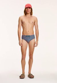 Sinised ja punased ujumispüksid mustrilise kangaga, millel on türkiissinine pael. Modell kannab pruune sandaale ja roosat mütsi.