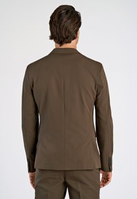 Ein Mann, von hinten betrachtet, trägt ein tailliertes braunes Sakko und passende Hose vor einem schlichten hellen Hintergrund.