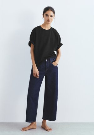 Donna in piedi a piedi nudi, indossa una maglietta nera larga con le maniche arrotolate e jeans blu scuro a gamba larga, su uno sfondo semplice.