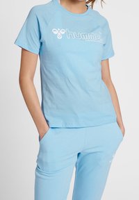Jasnoniebieska bawełniana koszulka z krótkim rękawem, okrągłym dekoltem i białym nadrukiem logo z przodu. Gładka tekstura, luźny krój.