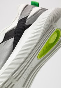 Baskets en cuir et textile gris avec une sangle noire, semelle blanche et languette de talon verte. Caractéristiques de la semelle rembourrée et détail de la semelle intérieure ronde et vert vif.