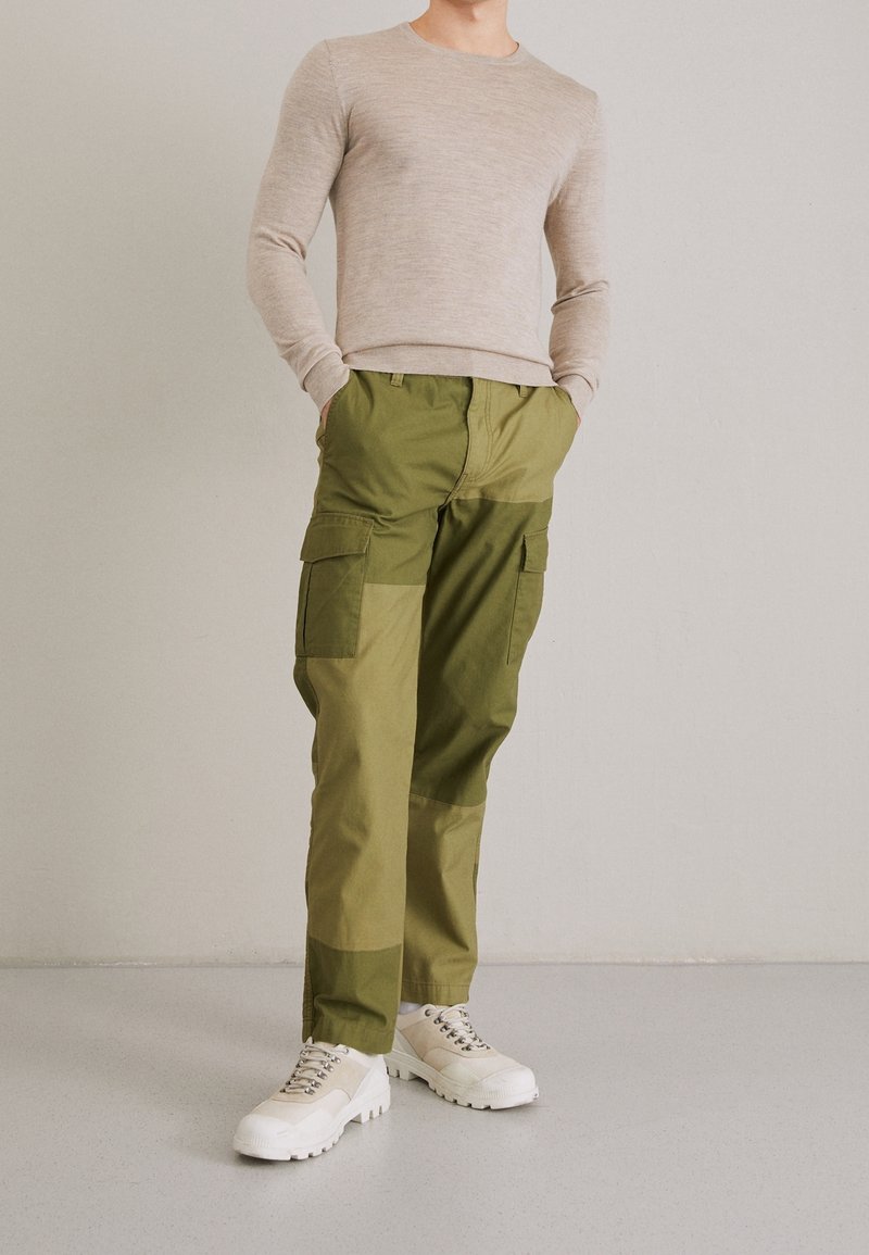 Personne portant une chemise beige à manches longues, un pantalon cargo vert bicolore et des baskets blanches épaisses, debout devant un mur clair uni.