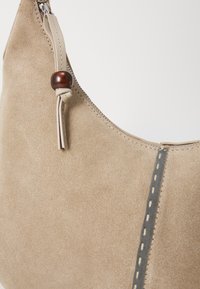 Beige mokkanahkakäsilaukku, jossa on vetoketjukiinnitys, ja jonka sivussa on puuhelmiakseli sekä kontrastivärinen harmaa tikkaus.