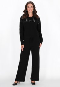 Fekete kötött pulóver V-nyakkal és díszes flitteres részletekkel a vállaknál, széles szárú fekete nadrággal és magas sarkú cipővel párosítva.