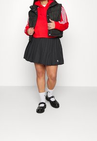 Svart puffig väst över en röd hoodie, tillsammans med en plisserad svart kjol, vita strumpor och svarta Mary Jane-skor med spännen.