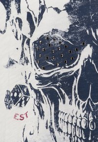 Biała koszulka z niebieskim graficznym motywem czaszki, ozdobiona srebrnymi ćwiekami, z nadrukiem kostki obok tekstu "EST" w kolorze czerwonym.