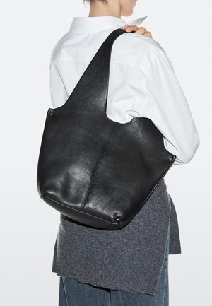 Persoon die een wit overhemd en een grijze rok draagt, houdt een grote zwarte leren tas over de schouder tegen een witte achtergrond.