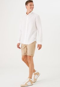 Weißes Leinen-Hemd mit Knöpfen,beige Shorts und hochgeschlossene Sneaker mit beigen Akzenten. Modell läuft und zeigt lässige Sommerkleidung.