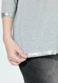 Eine Hand hält den Saum eines hellgrauen Shirts mit silbernen Streifen, kombiniert mit schwarzen Hosen.