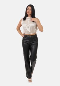 Donna con i capelli neri lisci che indossa una blusa beige lucida senza maniche, pantaloni in pelle nera e sandali bianchi con le punte scoperte, in posa su uno sfondo bianco.