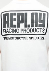 Λευκό πουκάμισο με μεγάλο μαύρο κείμενο "REPLAY RACING PRODUCTS" και μικρότερο κείμενο από κάτω που γράφει "THE MOTORCYCLE SPECIALISTS".
