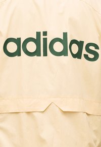 Tissu beige avec un grand texte de marque "adidas" vert foncé imprimé sur toute la surface, avec un petit détail en forme d'encoche triangulaire sous le texte.