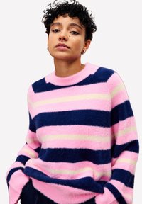 Personne avec des cheveux courts et bouclés portant un pull en laine rayé rose, bleu marine et crème, se tenant avec les mains dans les poches devant un fond blanc.