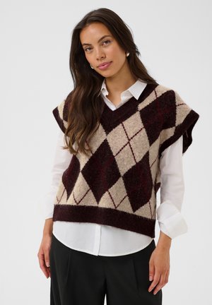 Femme aux longs cheveux bruns portant des boucles d’oreilles en perles, une chemise blanche et un gilet sans manches à motif losanges beige et brun foncé.