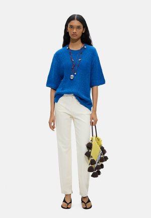 Kobieta ubrana w niebieski sweter z krótkim rękawem, białe spodnie, czarne sandały, warstwowe naszyjniki, trzymająca żółtą torbę z ciemnymi frędzlami.