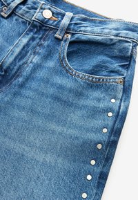 Jean en denim bleu en gros plan montrant la poche avant, la braguette à boutons, les passants de ceinture, et des clous argentés décoratifs le long de la couture latérale.