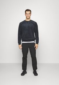 Mies seisoo kasvoillaan eteenpäin, yllään musta huppari, mustat housut, mustat lenkkarit ja valkoinen aluspaita vaaleaa taustaa vasten.