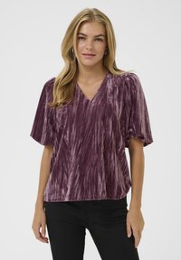 Lila Samtbluse mit V-Ausschnitt, kurzen Puffärmeln und einem strukturierten Streifenmuster. Leicht lockere Passform, kombiniert mit schwarzen Hosen.