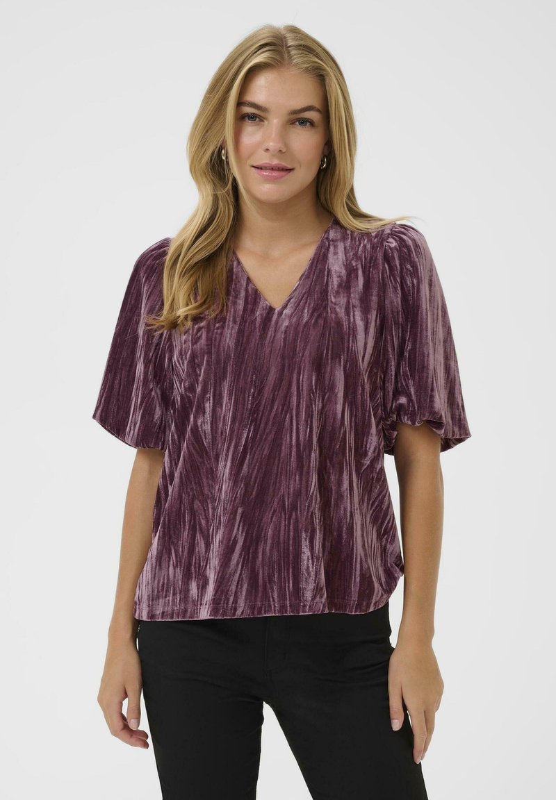 Lila Samtbluse mit V-Ausschnitt, kurzen Puffärmeln und einem strukturierten Streifenmuster. Leicht lockere Passform, kombiniert mit schwarzen Hosen.