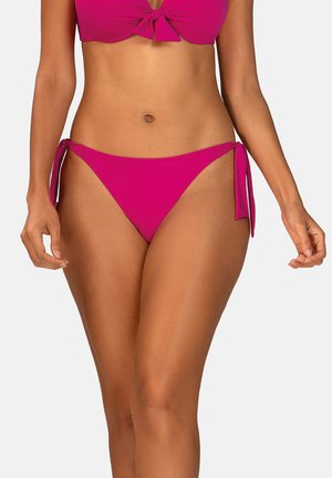 Kobieta w różowym bikini z wiązanymi po bokach majtkami i topem wiązanym z przodu, stojąca na tle jasnego, jednolitego tła.