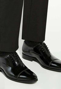 Glanzende zwarte formele schoenen met een cap-toe ontwerp, gedragen met zwarte broeken tegen een lichte achtergrond.