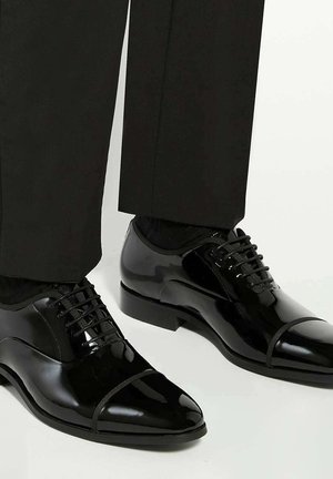 Shiny black formal dress shoes with cap-toe design worn with black trousers against a light background.