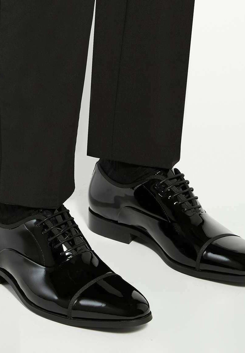 Glanzende zwarte formele schoenen met een cap-toe ontwerp, gedragen met zwarte broeken tegen een lichte achtergrond.