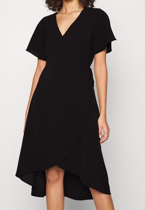 Frau trägt ein schwarzes Wickelkleid mit V-Ausschnitt, kurzen flatternden Ärmeln und asymmetrischem Saum, der über und unter den Knien endet.