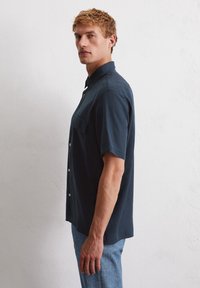 Koszula z krótkim rękawem w ciemnoniebieskim materiale z guzikiem z przodu, pojedynczą kieszenią na piersi oraz luźnym krojem o minimalistycznym designie.
