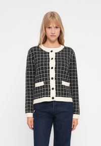 Μαύρη και λευκή καρό ζακέτα με ριχτές κρεμ λεπτομέρειες, στρογγυλή λαιμόκοψη, κουμπωτή μπροστά και δύο παPatch τσέπες. Μαλακή πλέξη.